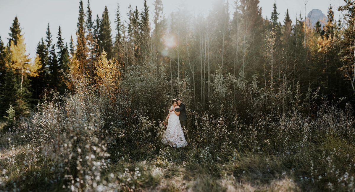 A couple in wedding attire embraces in a sunlit forest clearing, surrounded by autumn foliage, as their photographer captures the intimate elopement amid tall trees and golden light.