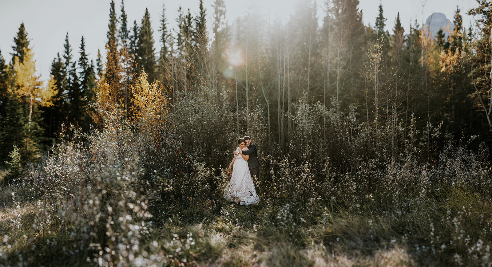 A couple in wedding attire embraces in a sunlit forest clearing, surrounded by autumn foliage, as their photographer captures the intimate elopement amid tall trees and golden light.