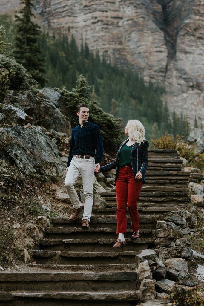 A man and woman hold hands while walking down stone steps outdoors in Banff, surrounded by pine trees and rocky mountains, both smiling at each other as their photographer captures this beautiful wedding moment.