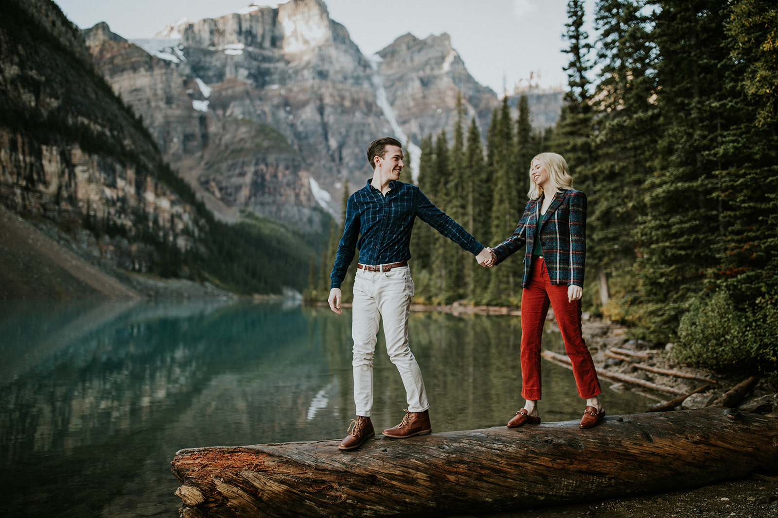 A couple holds hands on a large log by a mountain lake near Canmore, surrounded by pine trees and rocky peaks. Both smile in plaid shirts and boots—an adventurous elopement captured perfectly by their photographer.