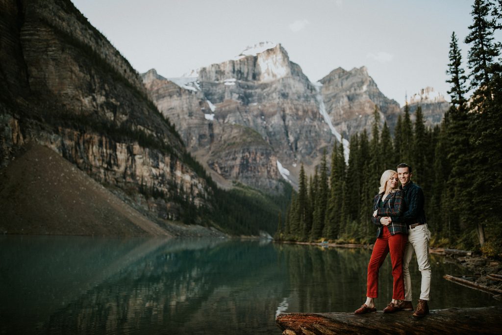 A couple stands on a log by a calm mountain lake near Banff, surrounded by pine trees and tall, rocky, snow-capped mountains in the background under a clear sky—perfect for an intimate elopement.
