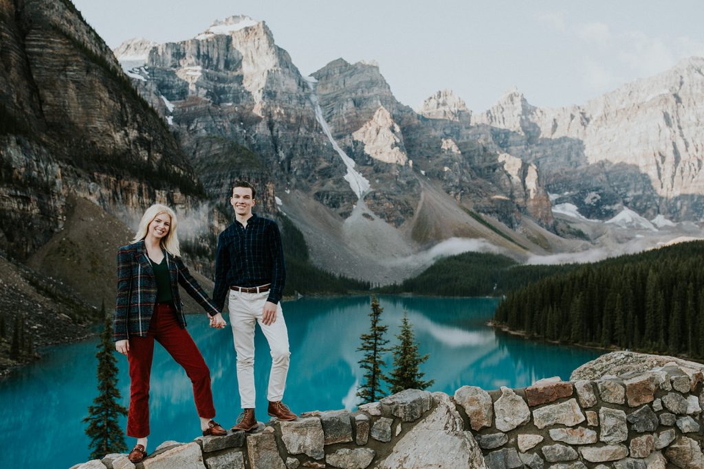 A smiling couple stands on a stone wall overlooking a turquoise lake, surrounded by tall pine trees and dramatic, snow-capped mountains under a clear sky—an unforgettable moment captured by a Canmore photographer.