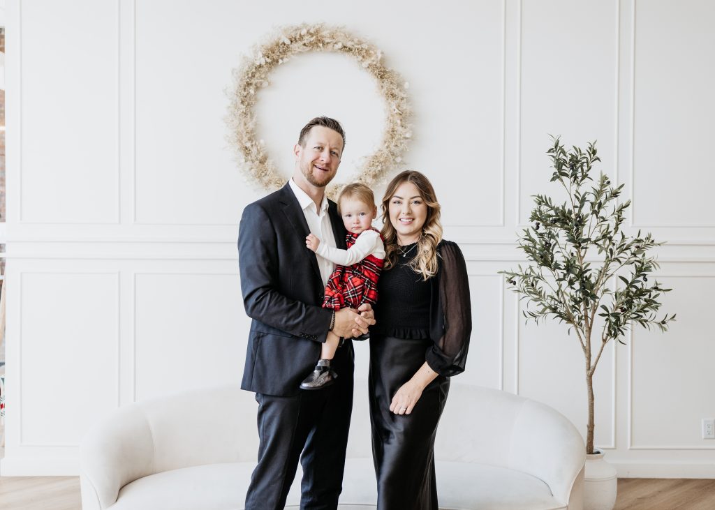 A man and woman stand smiling with a young child in festive attire, posing in front of a white sofa. A round wreath and small potted tree decorate the bright, elegant Calgary room.