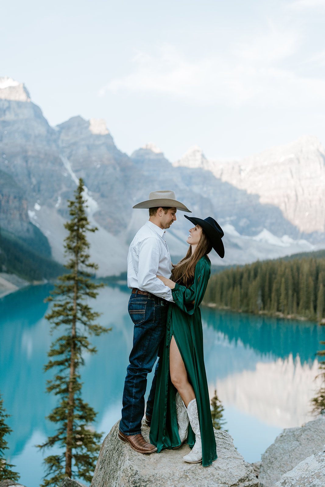 A couple in cowboy hats stands on a rock by a blue mountain lake near Canmore, surrounded by pine trees and rugged peaks. The woman wears a green dress and white boots as they gaze at each other, smiling on their wedding day.