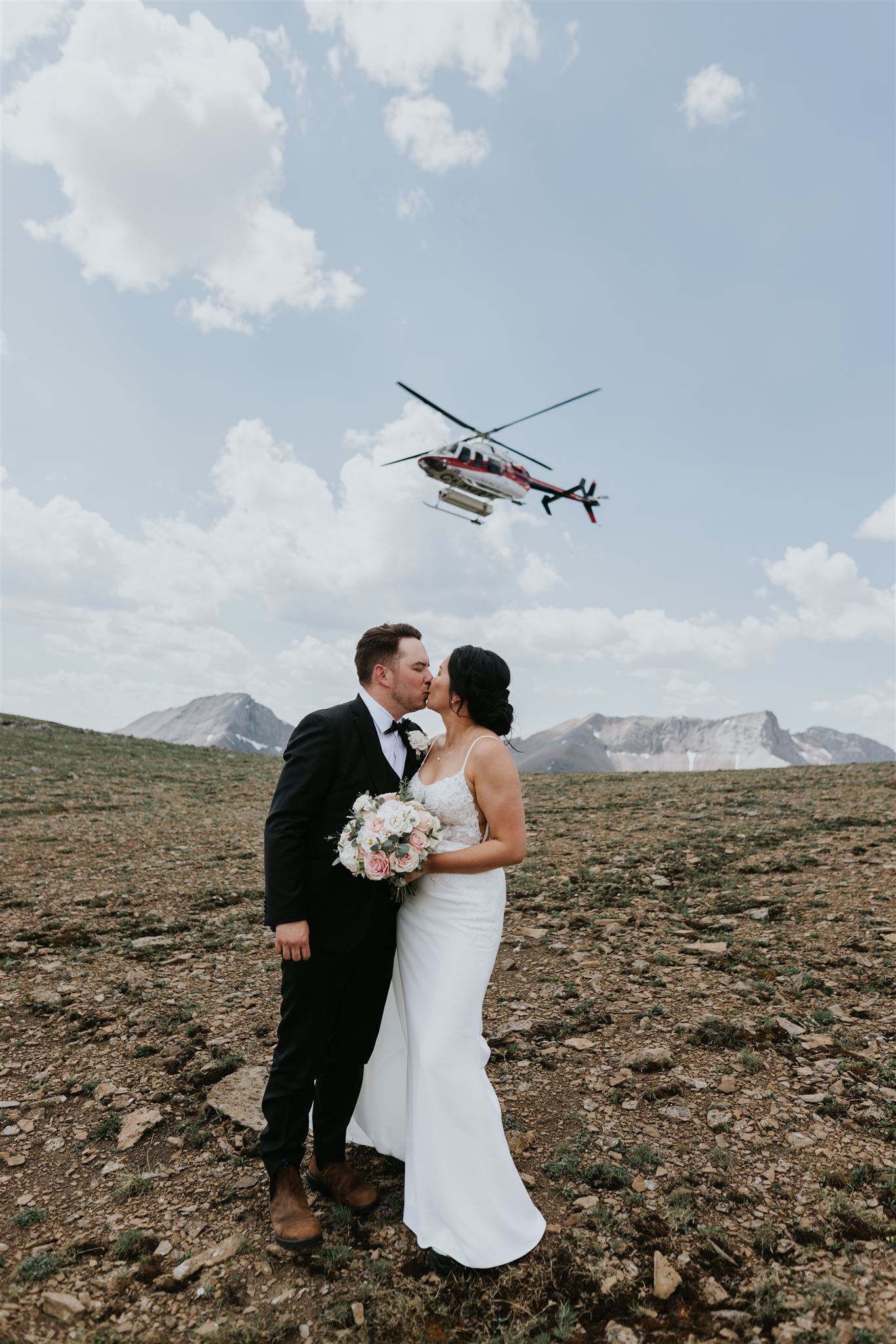A Canmore wedding unfolds as a bride and groom kiss on rocky terrain, mountains rising behind them. The groom in boots and a black suit, the bride in white with her bouquet—captured perfectly by a talented photographer as a helicopter passes overhead.