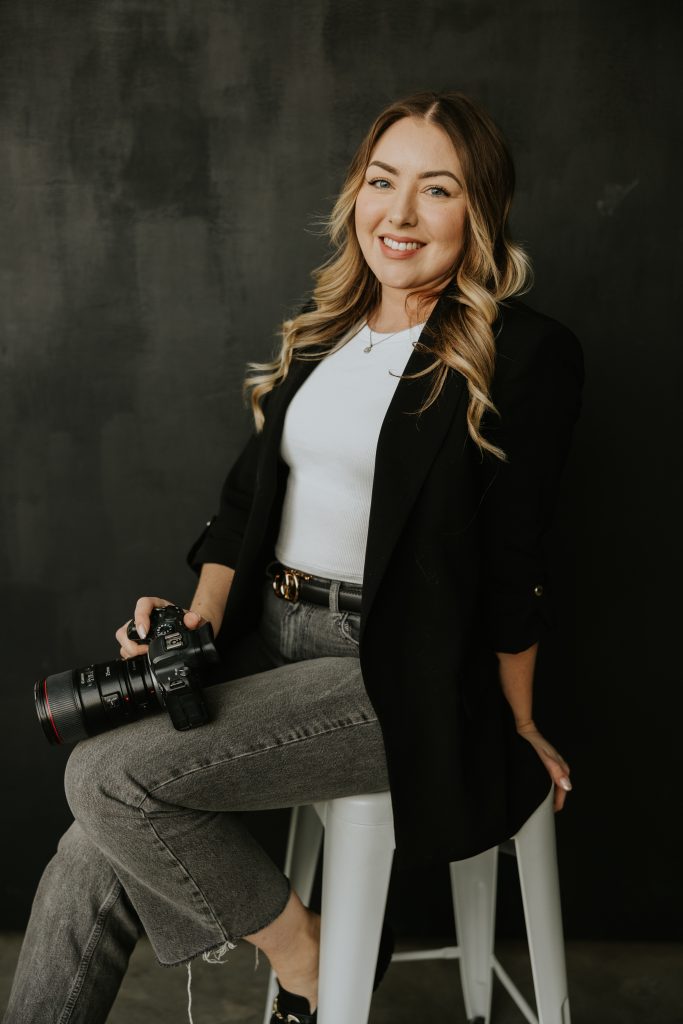 A woman with long, wavy blonde hair sits on a white stool against a dark background, smiling. Dressed in a black blazer and gray jeans, she holds a camera—ready to capture an elopement in Canmore or Banff.