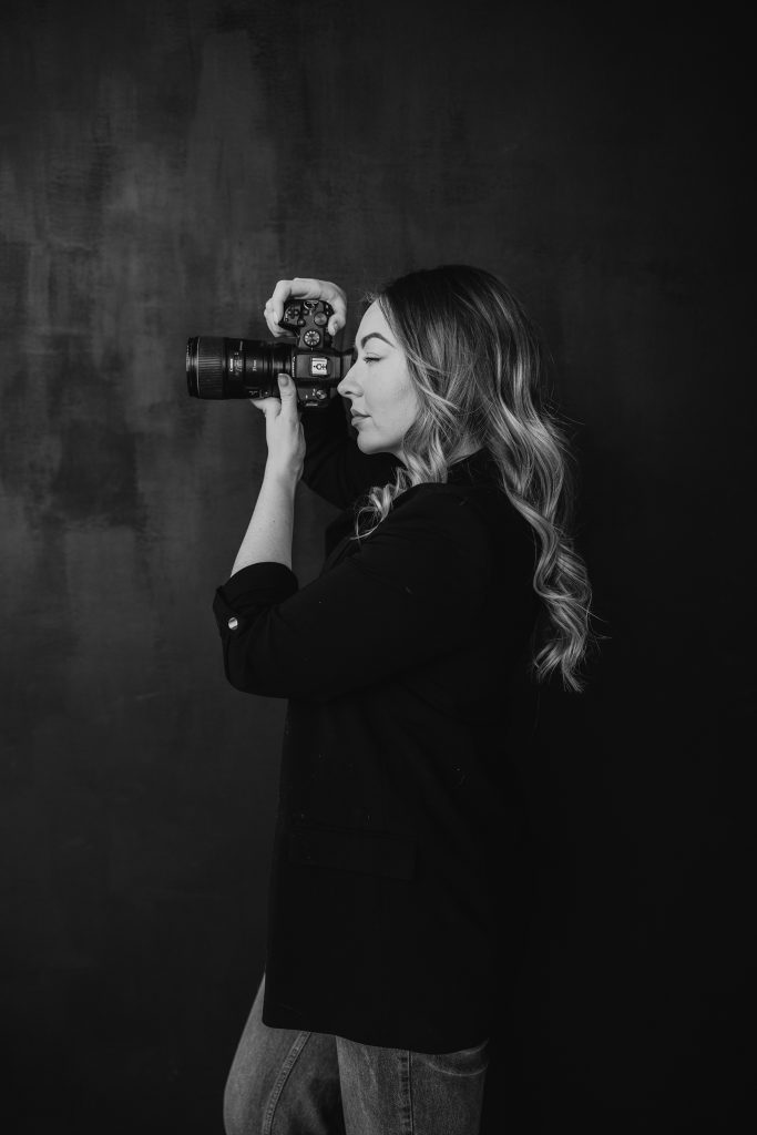 A woman with wavy hair holds a camera up to her eye, taking a photo against a dark, plain background. Shown in profile in black and white, she wears a dark blazer and jeans—capturing the spirit of a Canmore elopement.