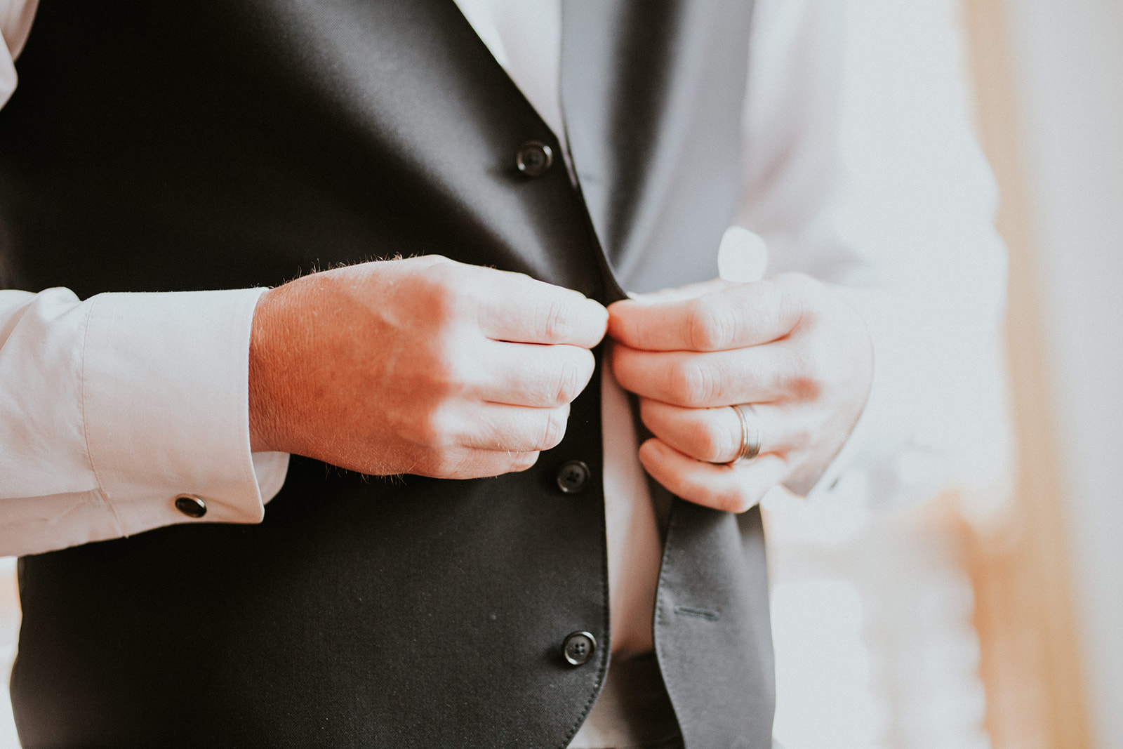 A person wearing a white dress shirt and dark vest, possibly preparing for a Banff elopement, buttons up the vest. Only their torso and hands are visible, with a wedding ring on their left hand.