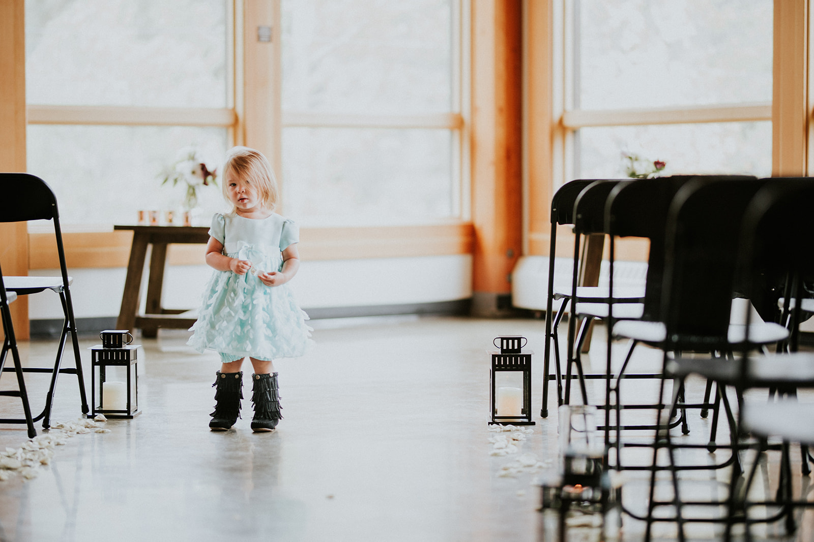 A young girl in a light blue dress and black boots walks down an aisle lined with black chairs and lanterns, scattered with white flower petals, at a beautiful Banff wedding in a bright room with large windows.