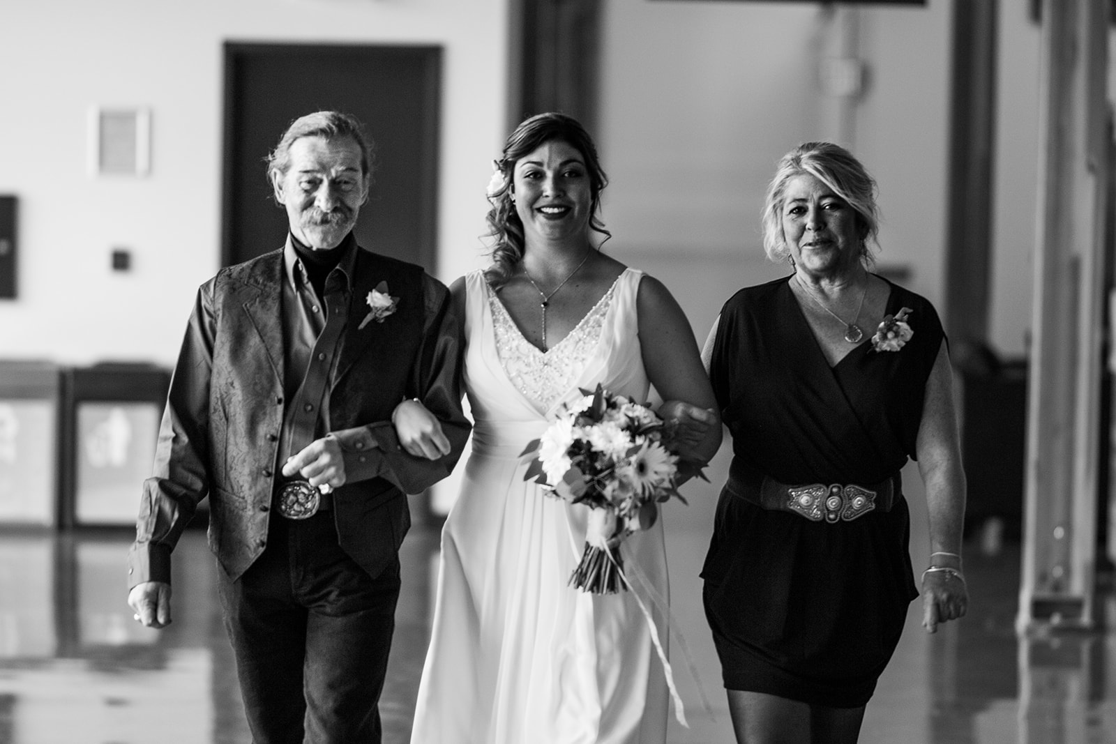 A bride in a white dress, holding a bouquet, walks arm in arm with an older man and woman, all smiling as they move forward together in a brightly lit indoor space—captured beautifully by their Banff wedding photographer.