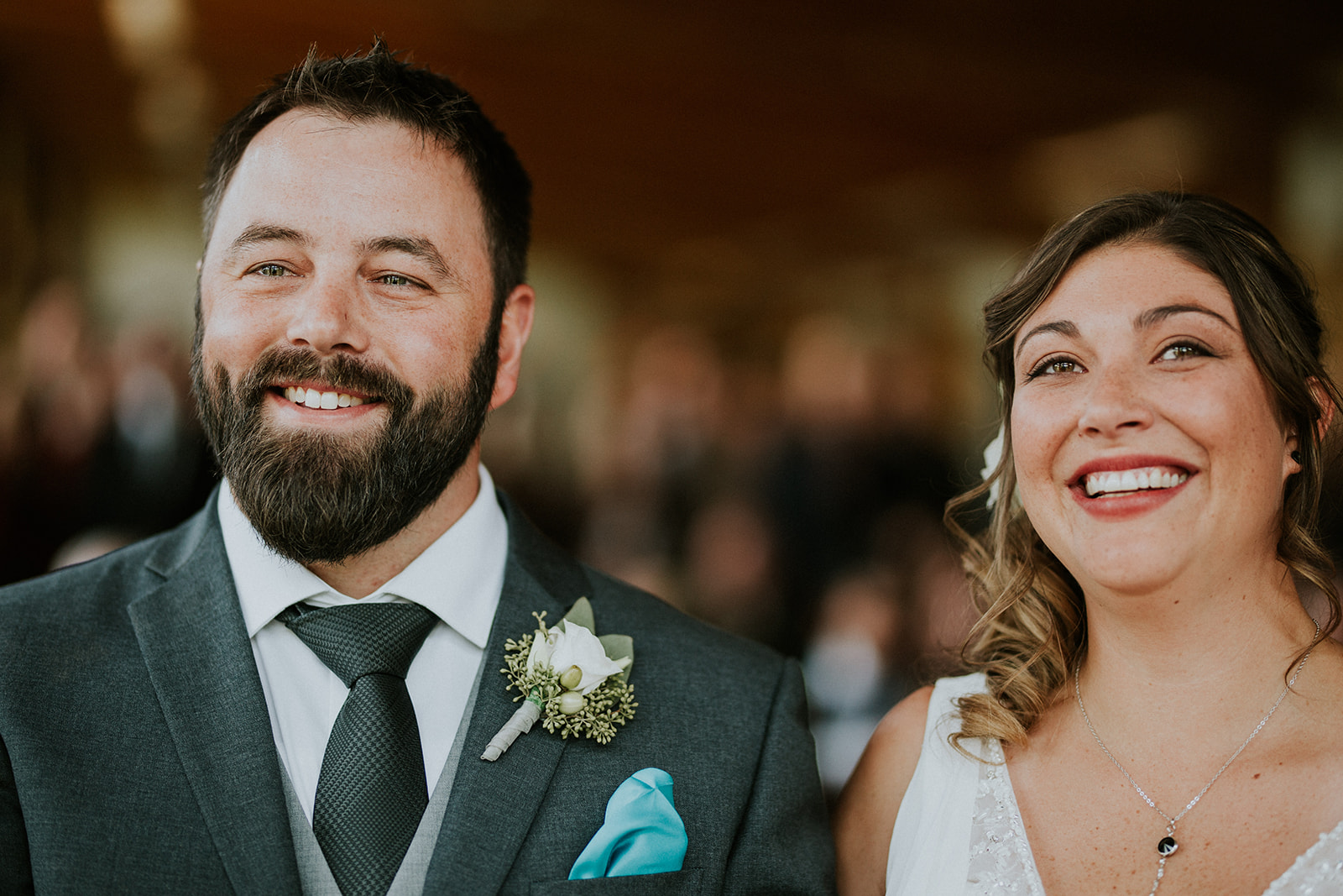 A man in a suit with a boutonnière and a woman in a white dress and necklace smile happily side by side, likely at an intimate elopement, with a blurred crowd in the background.