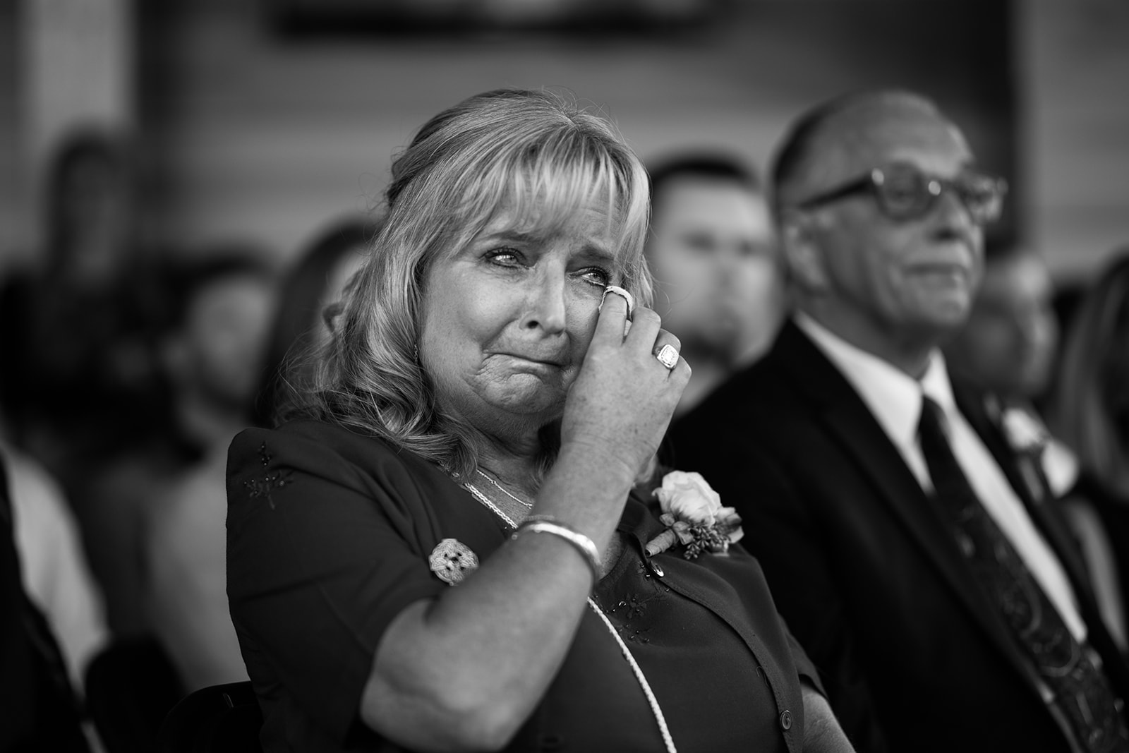 A woman wipes away tears while sitting in an audience at a formal wedding, wearing a dress and corsage. A man in a suit and glasses sits beside her. The black and white image captures the emotion, skillfully shot by a Banff photographer.