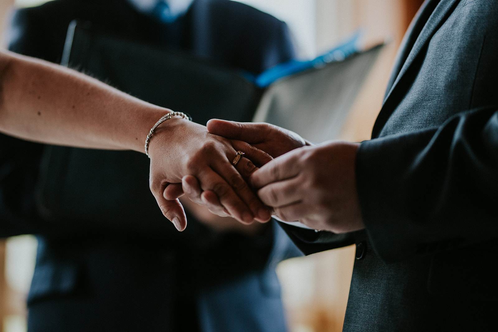 A close-up of two people exchanging rings during a wedding ceremony in Canmore, with one person gently placing a ring on the other's finger while a photographer and a figure in a suit holding a folder appear in the background.