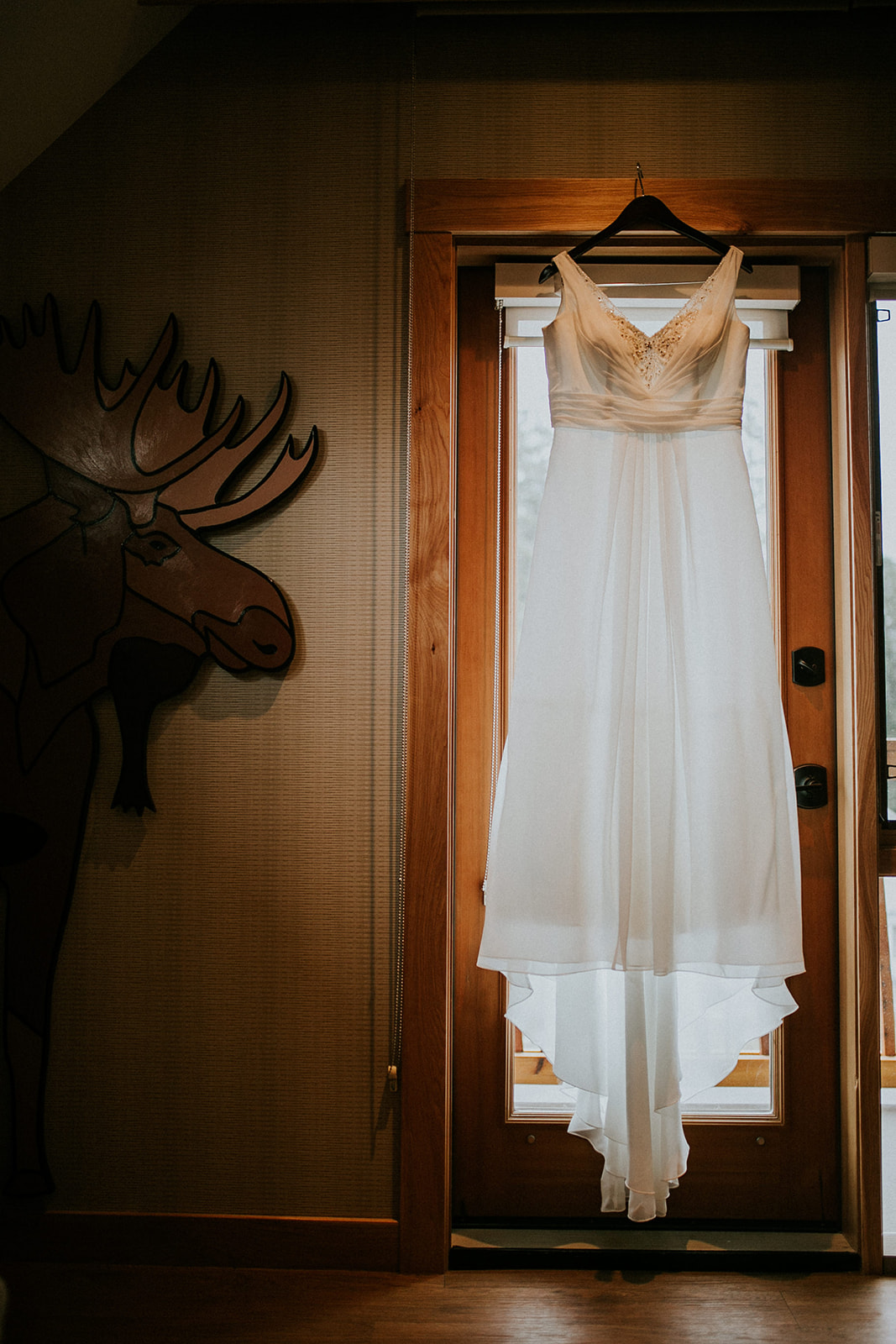 A white wedding dress hangs on a wooden hanger in front of a glass door, with soft natural light shining through—a beautiful scene for an intimate Calgary elopement. A painted moose is visible on the wall to the left of the door.