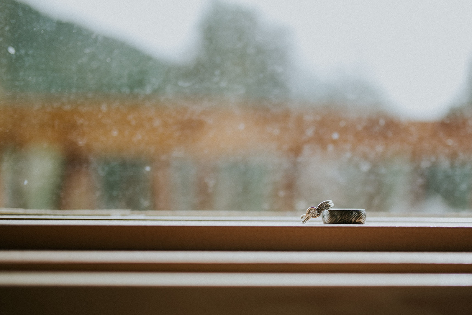 Two rings, one with a diamond, rest side by side on a wooden windowsill. The blurred window hints at trees beyond, captured perfectly by a photographer—an intimate scene ideal for an elopement in Banff.