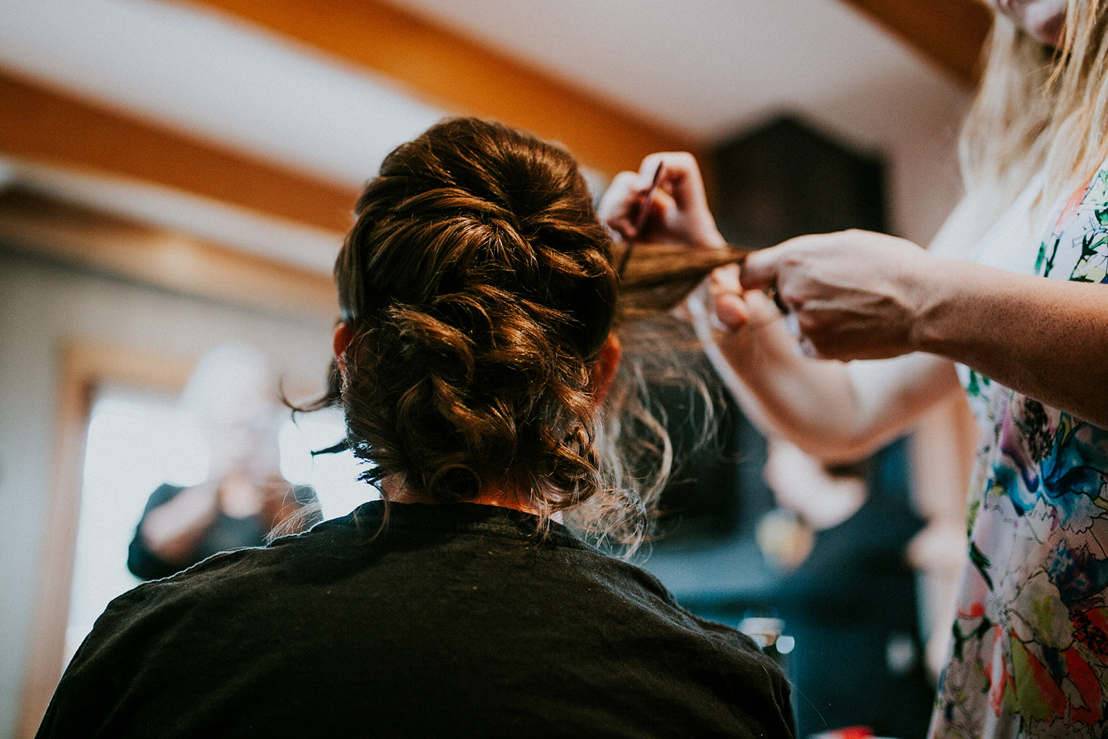 A person with brown hair sits facing away while another styles their hair, twisting and pinning sections up—capturing a serene elopement moment indoors in Canmore, with natural light and wooden ceiling beams above.