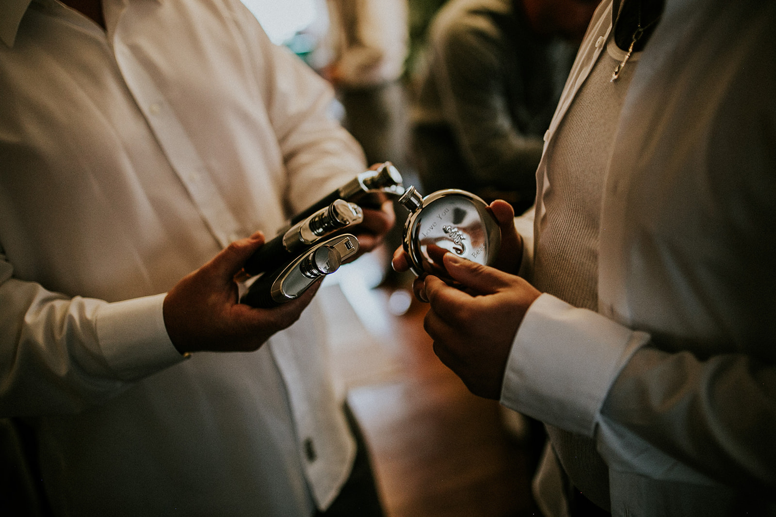 Two people in white dress shirts hold metal hip flasks and a pocket watch, preparing for a wedding. The focus is on their hands and the objects; their faces are out of frame, capturing the anticipation of a special day in Calgary.