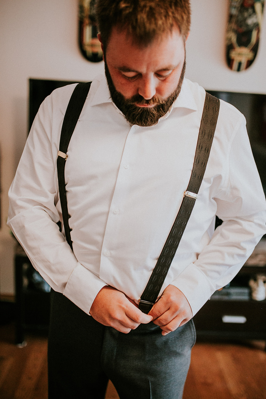 A bearded man wearing a white dress shirt and dark suspenders looks down as he adjusts his suspenders. Captured by a wedding photographer, he stands indoors on wooden floors with a TV in the background, evoking Canmore charm.