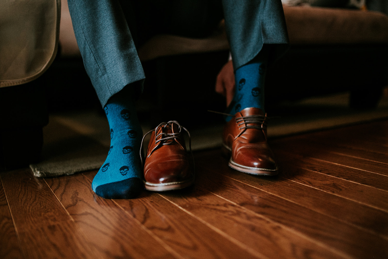 A photographer in blue dress pants sits on a sofa, wearing bright blue socks with black skull patterns and brown leather dress shoes—one shoe on, the other foot still in just a sock—on a wooden floor, perhaps prepping for a Banff or Canmore shoot.