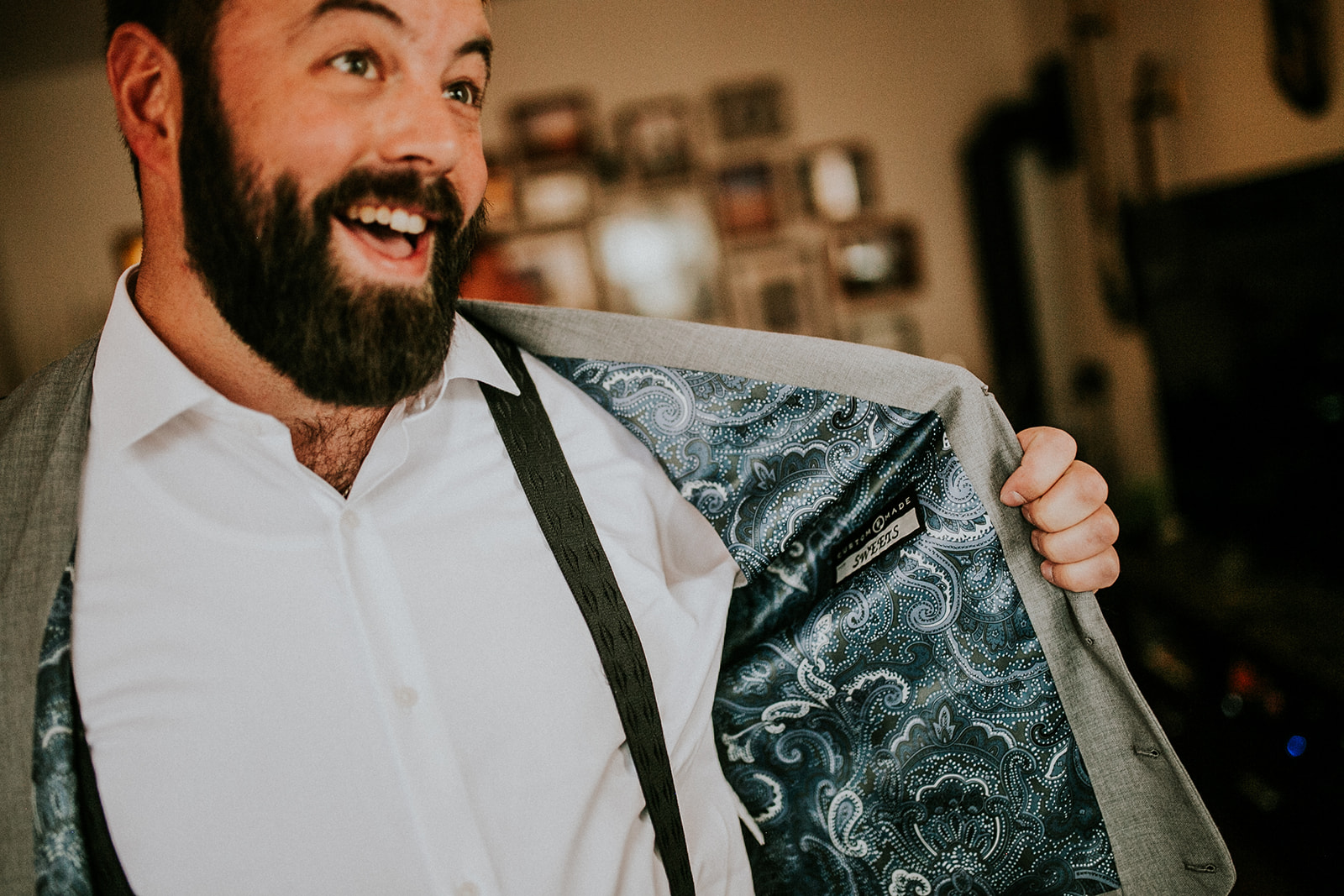 A bearded man smiles widely while holding open his grey suit jacket, revealing a blue paisley-patterned lining. Dressed for a wedding in Calgary, he wears a white shirt and suspenders, standing indoors with blurred photos on the wall behind him.
