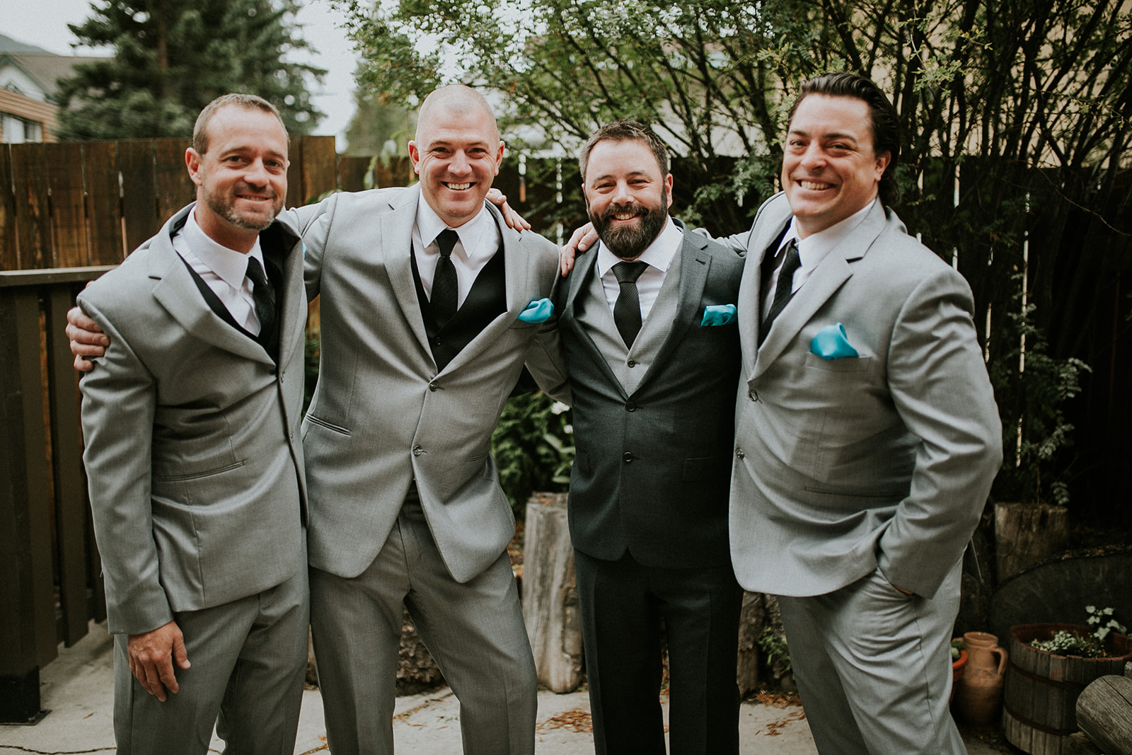 Four men dressed in suits, three in light grey and one in black, stand outdoors smiling with their arms around each other, posing for a group photo in a beautiful garden setting by a Calgary photographer.