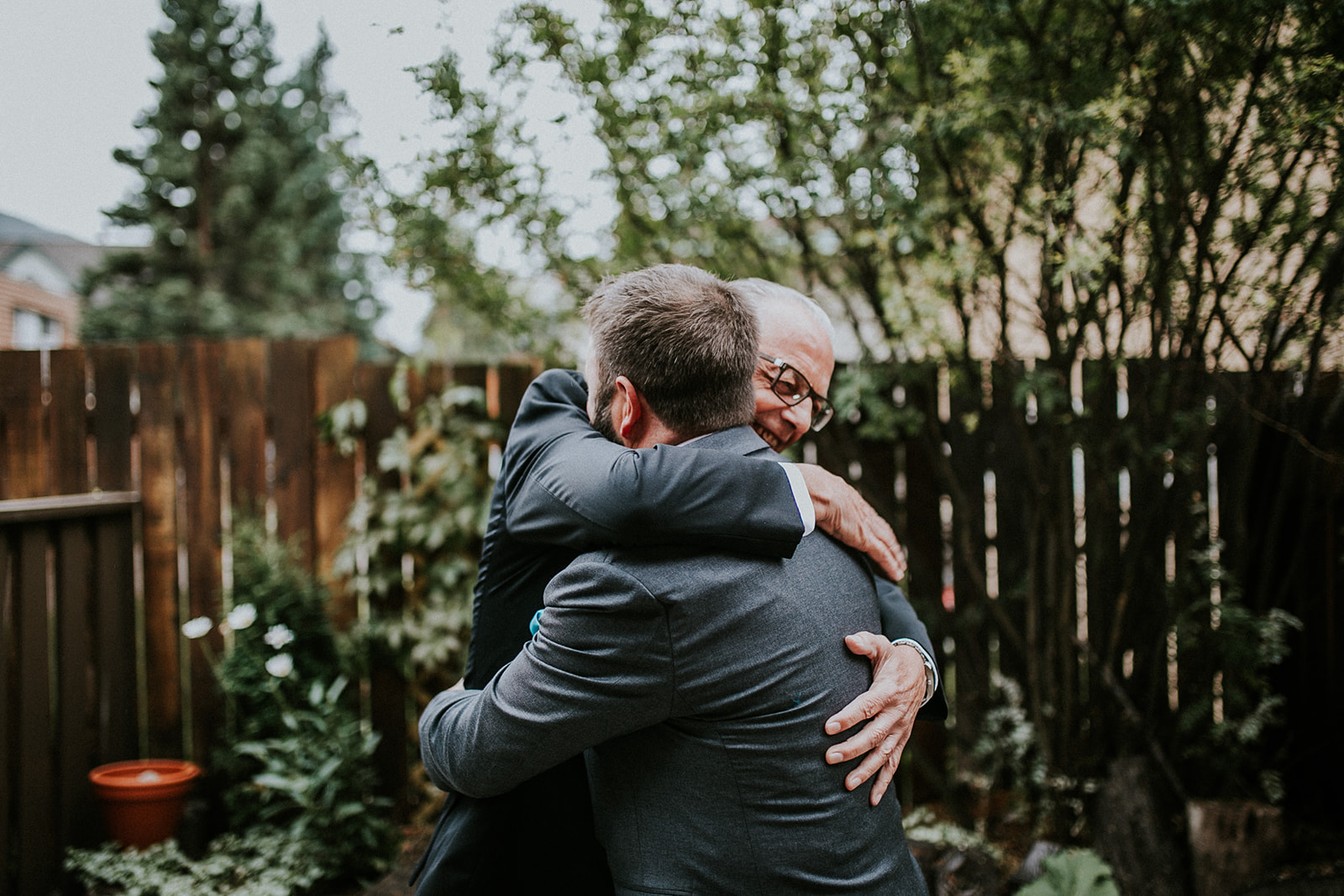 Two men in suits warmly embrace each other outside, surrounded by greenery and a wooden fence, both smiling with joy—captured perfectly by a photographer in Calgary.