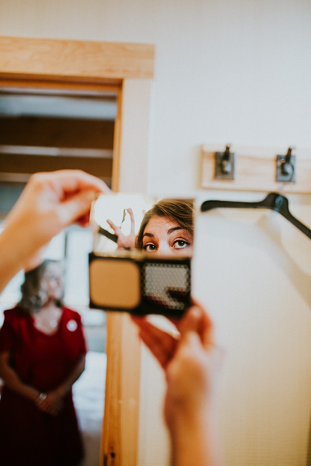 A person’s face is reflected in a compact mirror as they hold it up, focusing on their eyes. In the background, a woman in a red dress—perhaps ready for an elopement or wedding—stands blurred near a wooden doorway in Banff.