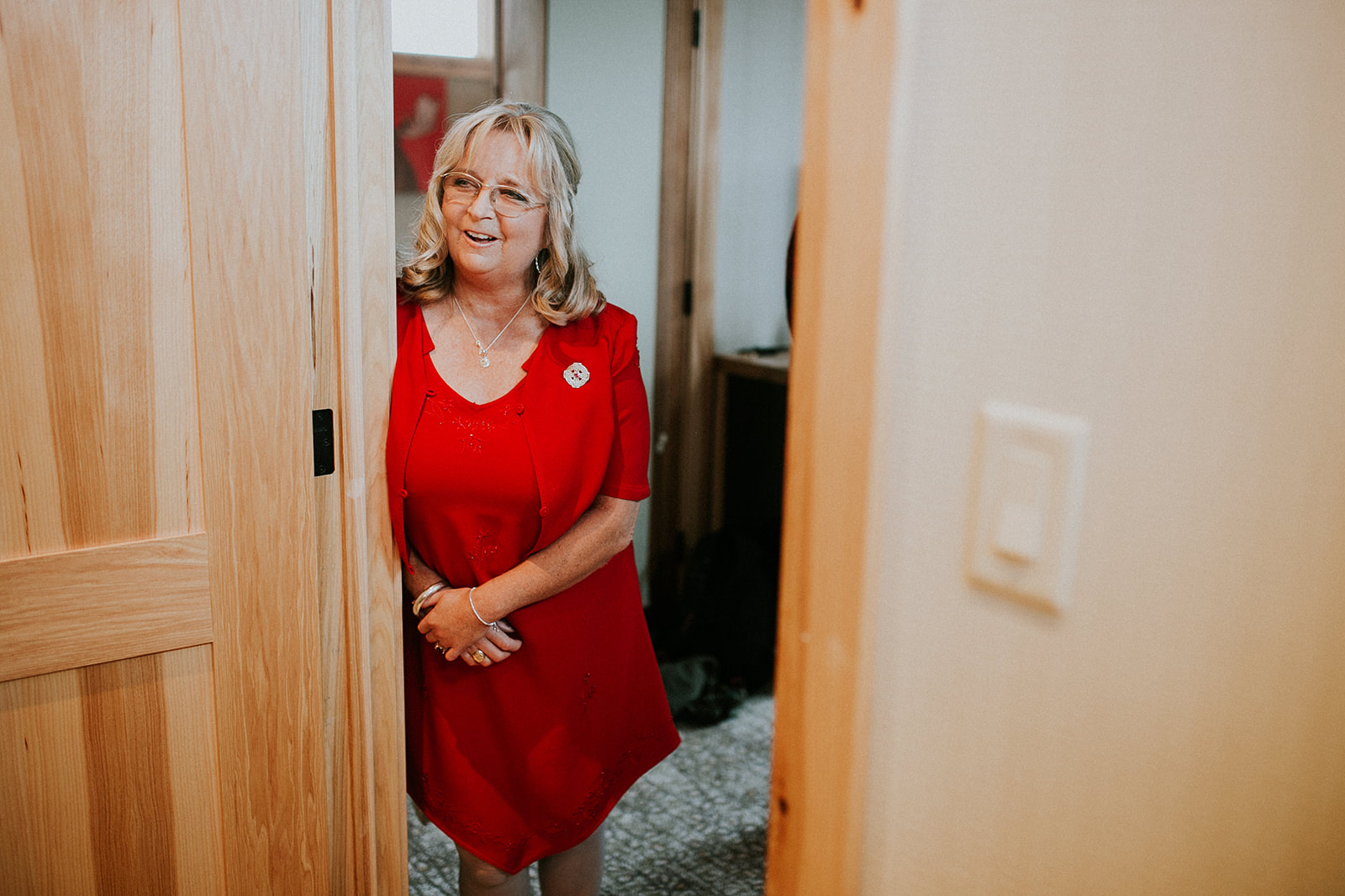 A smiling woman with blonde hair and glasses, wearing a red dress and a matching jacket, stands partially behind a wooden door in a warmly lit room, ready for her Calgary elopement.