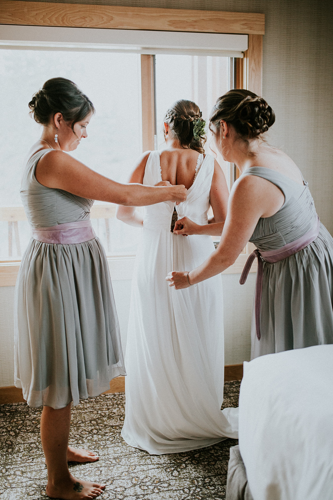 Two bridesmaids in light gray dresses with lavender sashes help a bride zip up her white wedding dress in a softly lit room near a window—perfect for an intimate elopement in Calgary, with the bride’s intricate updo and dress details on display.