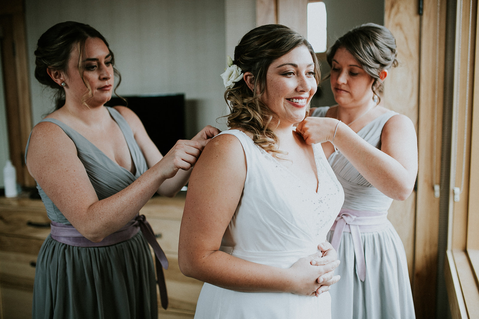 A bride in a white dress smiles as two bridesmaids in lavender-gray dresses help adjust her gown by the shoulders in a softly lit room with wooden furniture, beautifully captured by a Banff wedding photographer.