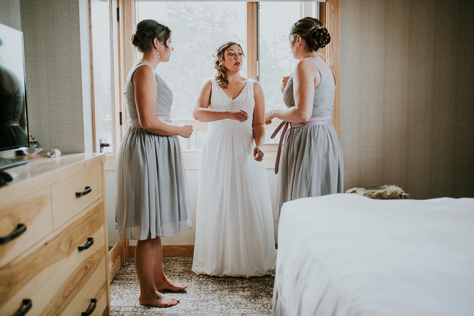 A bride in a white wedding dress stands between two bridesmaids in grey dresses, talking together by a window in a softly lit bedroom before their intimate Canmore elopement.