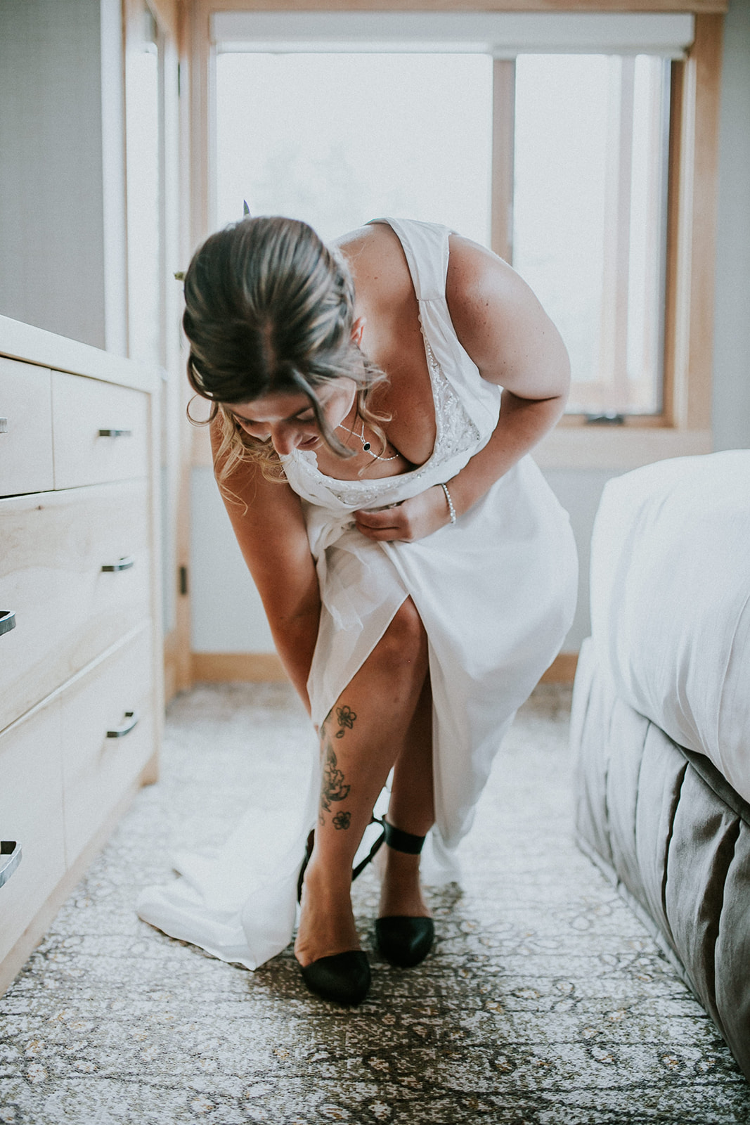 A woman in a white wedding dress bends down to fasten the ankle strap of her black shoe, revealing floral tattoos on her leg. A photographer captures this candid moment in a bright, cozy room with wood furnishings and a large window.