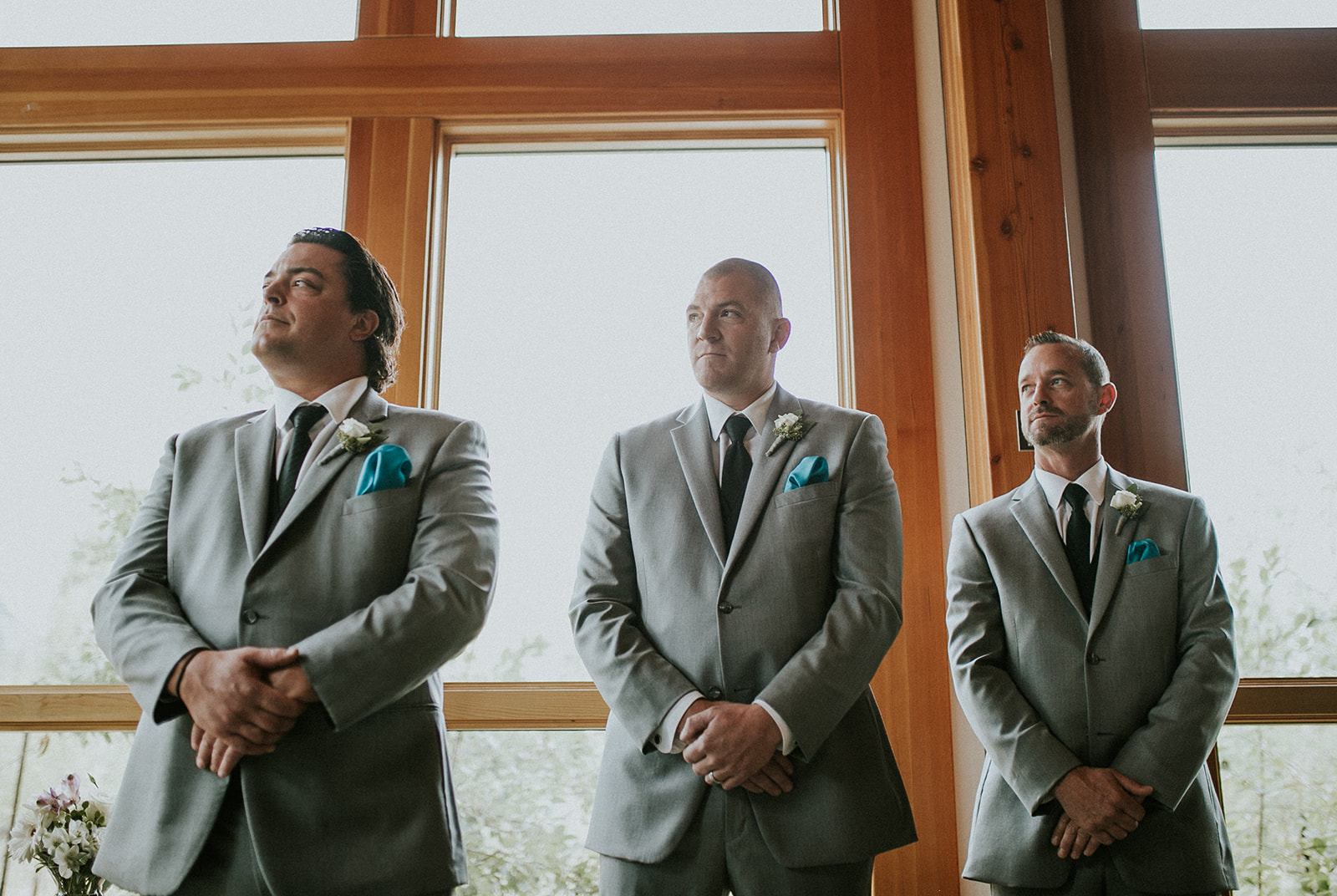 Three men in gray suits with turquoise pocket squares and white boutonnieres stand indoors in front of large windows, looking ahead with hands clasped, appearing to wait during a formal Banff elopement event.