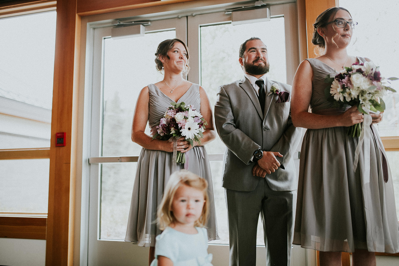 Three adults dressed formally, two women holding flower bouquets and one man in a suit, stand indoors by glass doors at a Banff wedding; a young girl in a light blue dress is blurred in the foreground.
