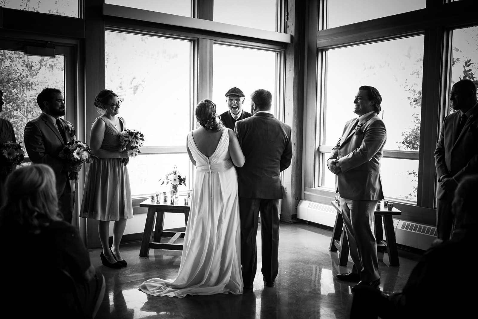 A black and white photo of a Calgary wedding ceremony indoors, with a bride and groom standing before an officiant. Bridesmaids and groomsmen gather as sunlight streams through large windows in the background.