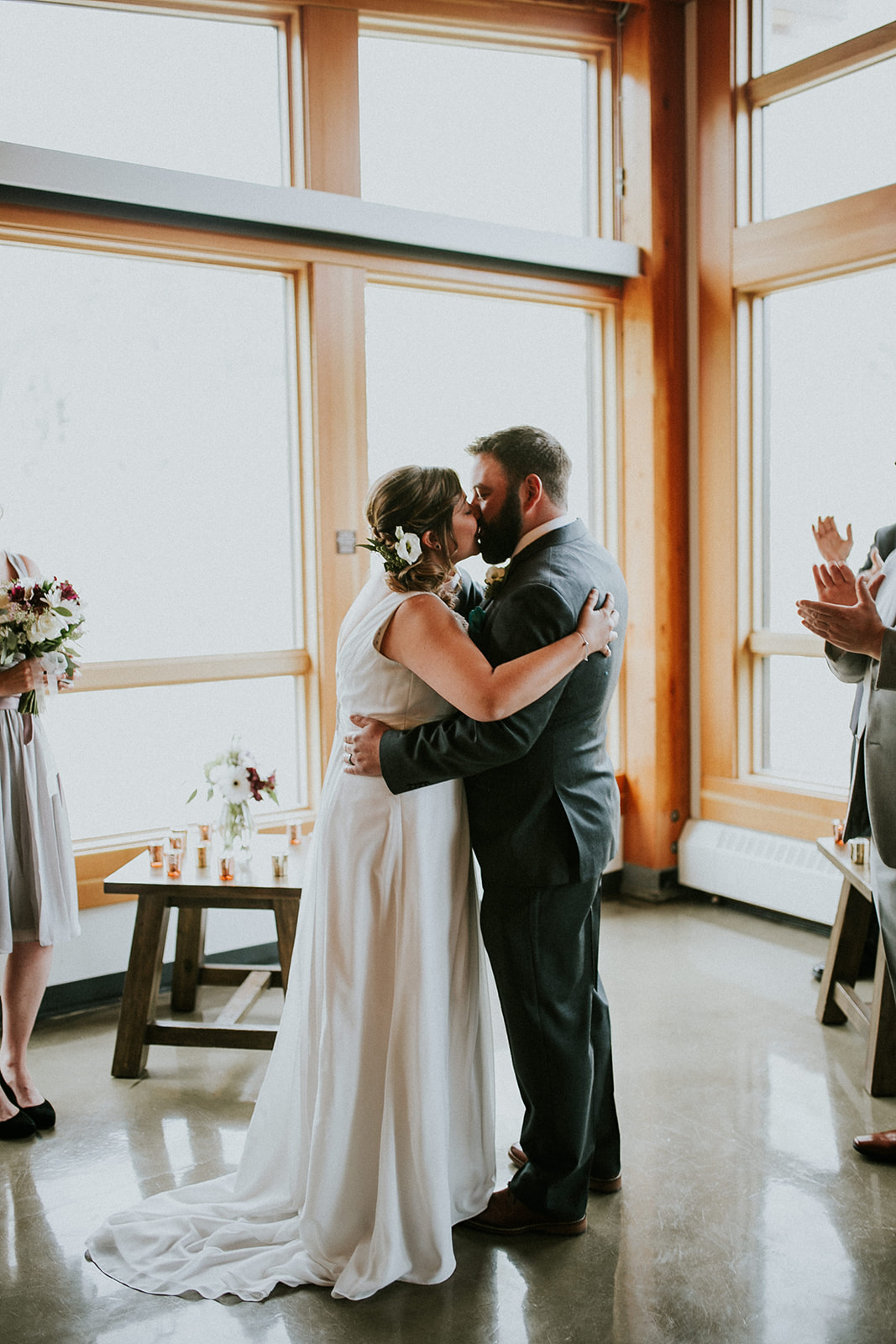 A bride and groom share a kiss indoors by large windows during their Banff wedding, with guests applauding nearby. The bride holds a bouquet in her hand, dressed in white, while the groom wears a suit.