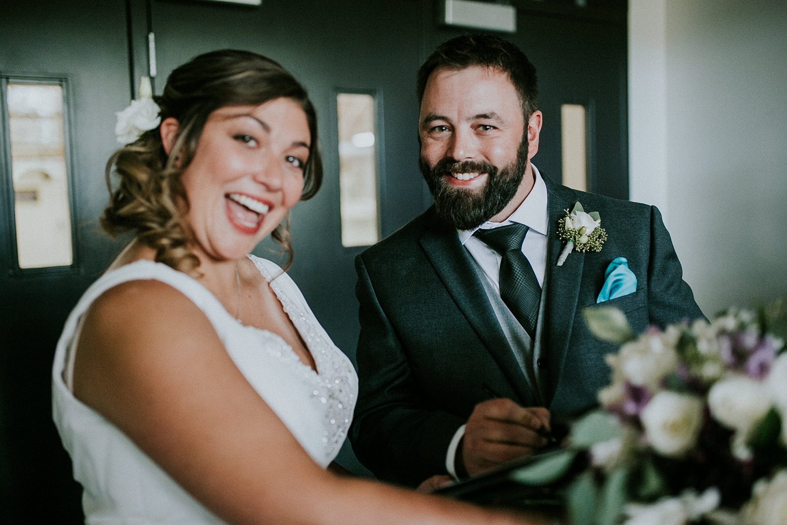 A smiling bride in a white dress and a groom in a dark suit pose happily indoors during their Calgary elopement, with the groom holding a pen and a bouquet of flowers visible in the foreground.