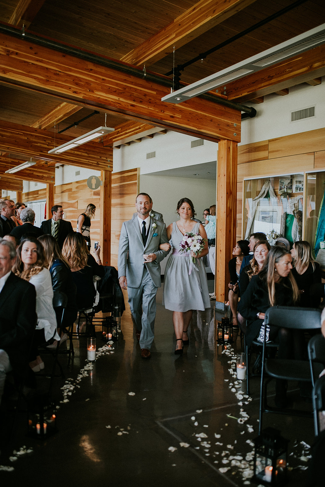 A man in a light grey suit and a woman in a light grey dress walk arm-in-arm down an aisle lined with seated guests, candles, and scattered flower petals at their intimate Canmore wedding in a warmly lit indoor venue with wooden beams.