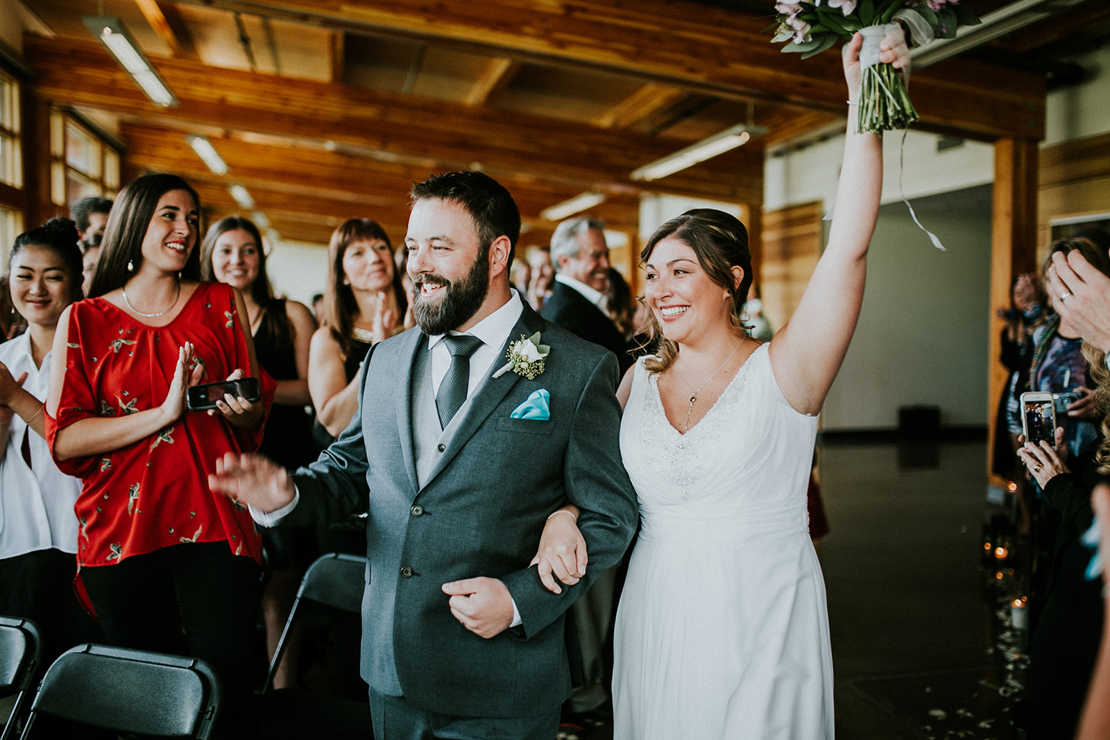 A smiling bride in a white dress holds up her bouquet while walking arm-in-arm with the groom, who wears a gray suit. Guests cheer and take photos during their Canmore elopement ceremony.