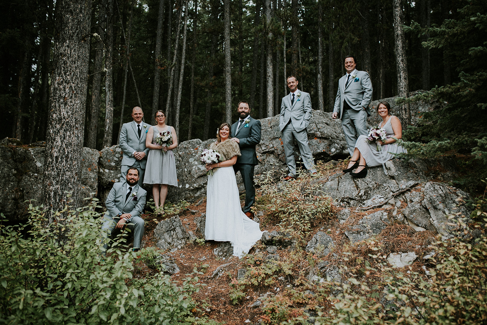 A wedding party poses outdoors on rocky Canmore terrain among trees; the bride in white and groom in grey stand at the center, surrounded by bridesmaids in grey dresses and groomsmen in grey suits, captured perfectly by their photographer.
