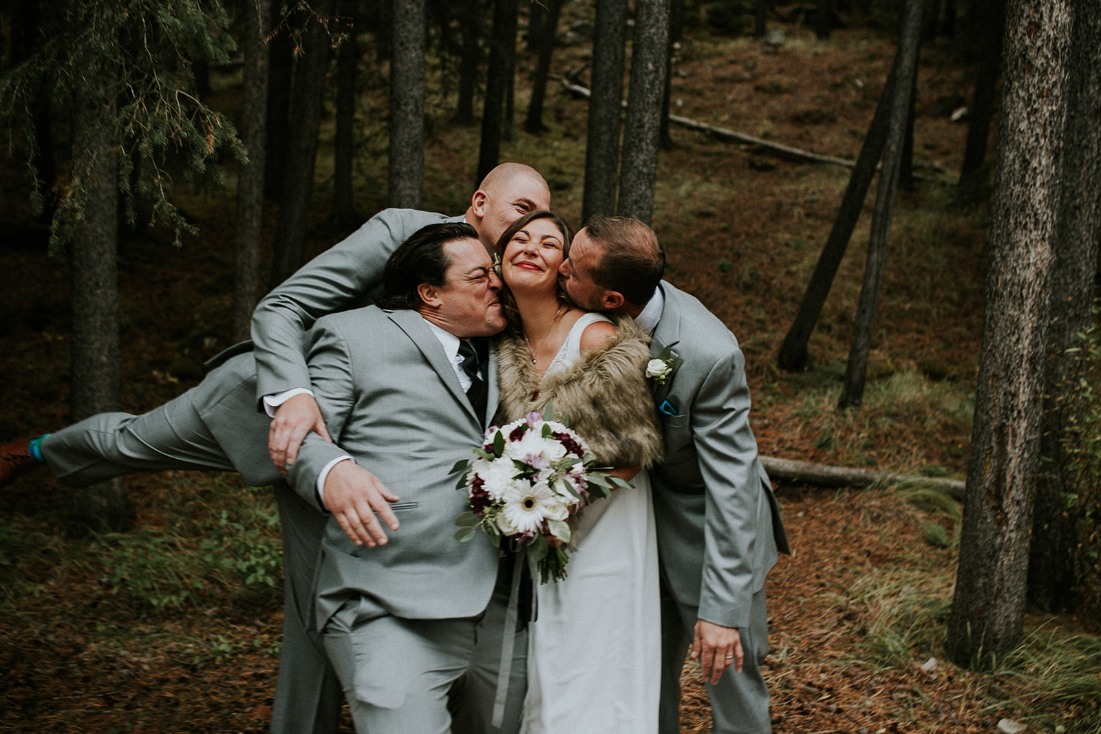 A bride in a white dress holding a bouquet is hugged and kissed on the cheeks by three groomsmen in grey suits, all laughing together during an elopement in a forested outdoor setting near Banff.