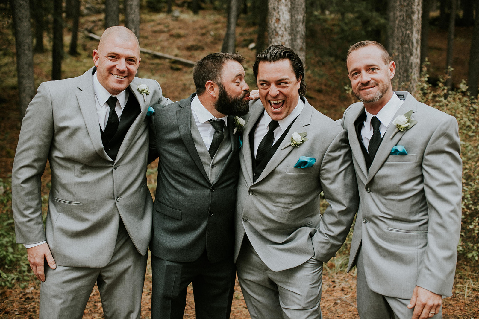 Four men in suits stand together in a forest near Canmore, smiling and laughing. Three wear light grey suits with teal pocket squares; the fourth wears a dark suit. They appear to be at a wedding or formal event.