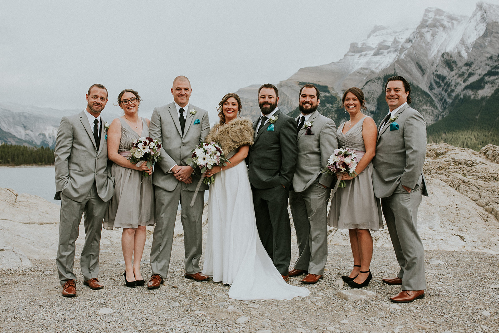 A wedding party poses outdoors by a lake and mountains in Banff. Captured by their photographer, the group of four men in grey suits and three women in grey dresses surrounds the smiling bride in her white gown and fur stole.