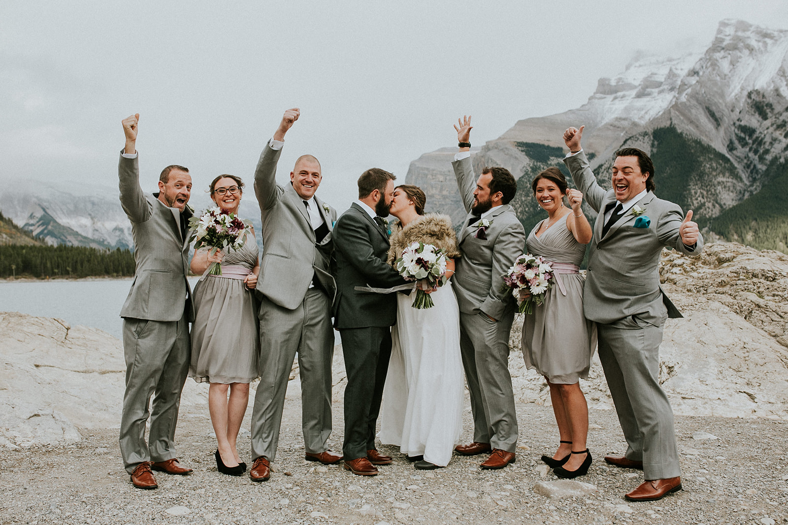 A wedding party celebrates outdoors in stunning Canmore, with rocky mountains and a lake as the backdrop. The bride and groom kiss in the center, surrounded by joyful bridesmaids and groomsmen, all captured by a talented photographer.