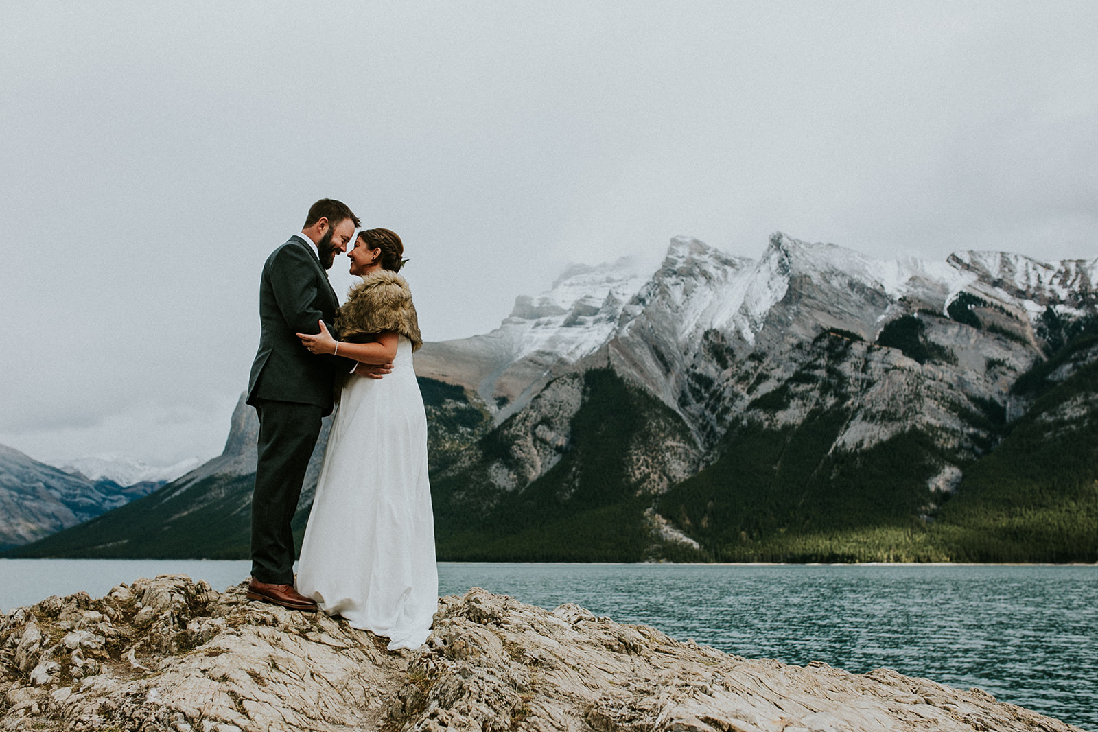 A bride and groom embrace on a rocky shore in Canmore, with a lake and snow-capped mountains behind them, captured by a photographer under a cloudy sky.