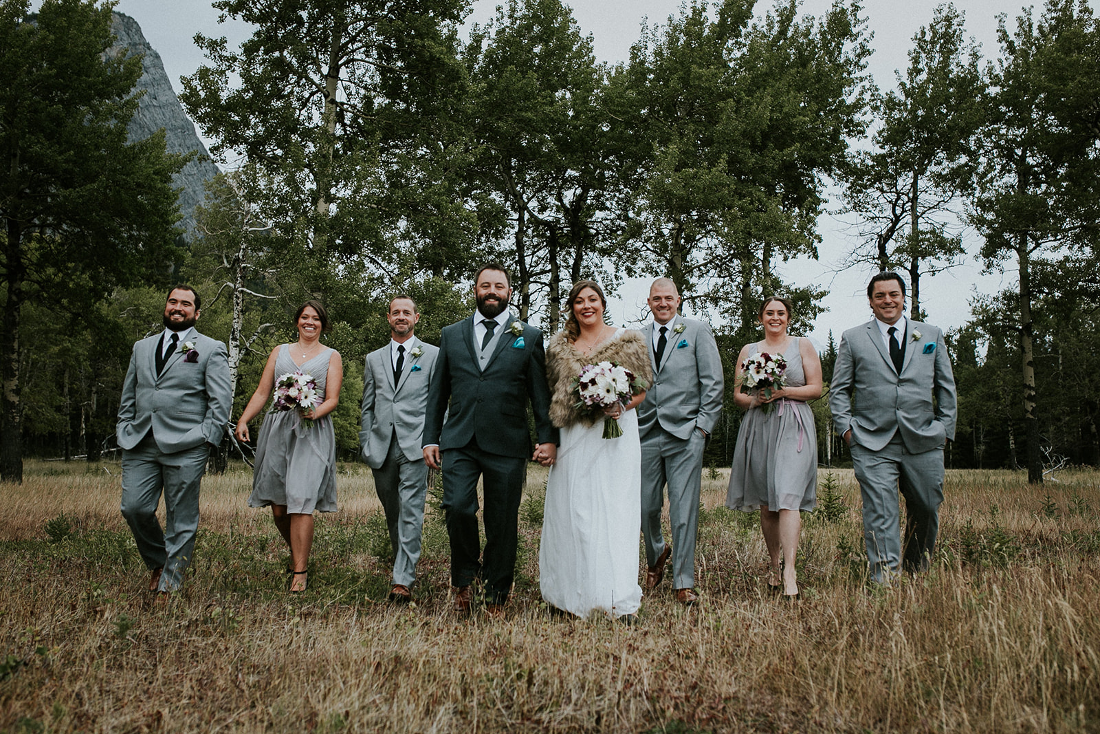 A wedding party walks outdoors in a grassy Canmore field, smiling. The bride in white holds a bouquet, flanked by the groom and attendants in grey suits and dresses, with trees and mountains in the background.