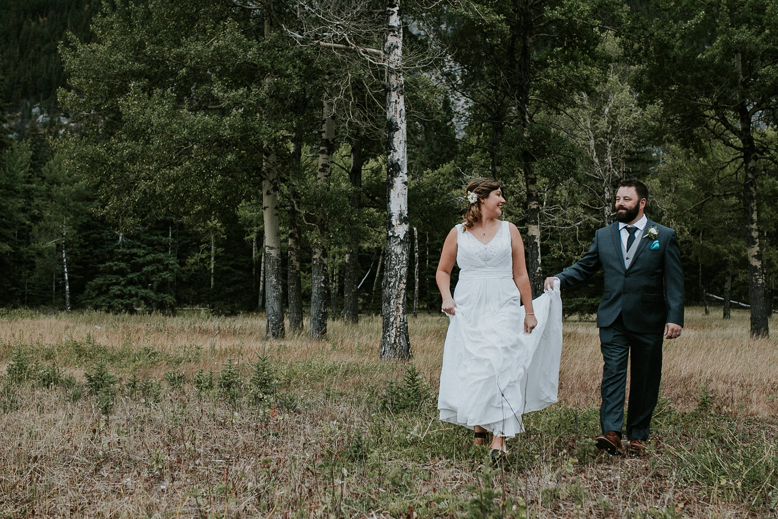 A bride in a white dress and a groom in a dark suit walk hand in hand through a grassy field surrounded by trees, smiling at each other on their wedding day, beautifully captured by a Calgary wedding photographer.