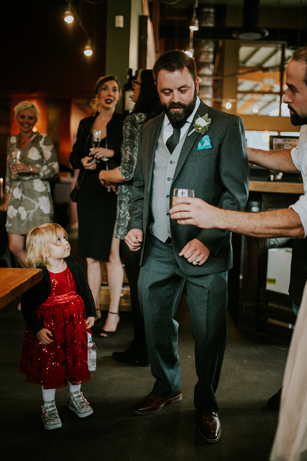 A man in a gray suit dances with a drink at a festive indoor wedding, as a young girl in a red dress watches. Several guests in semi-formal attire, also holding drinks and smiling, stand nearby—possibly celebrating in Canmore or Banff.