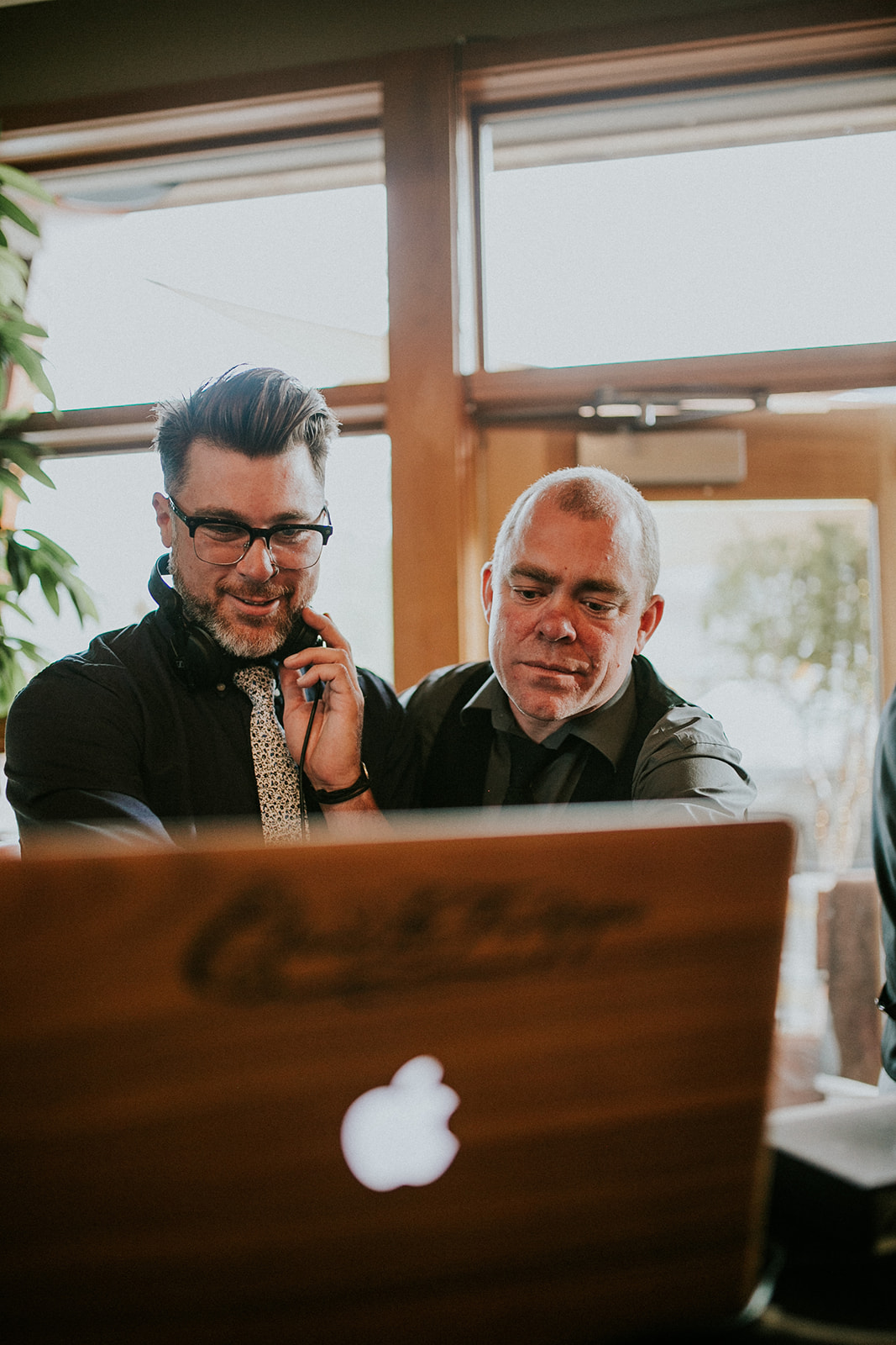 Two men dressed in black shirts stand together, looking at a laptop with an Apple logo. One wears glasses and headphones around his neck, while the other smiles as they work side by side in a warmly lit Calgary room.