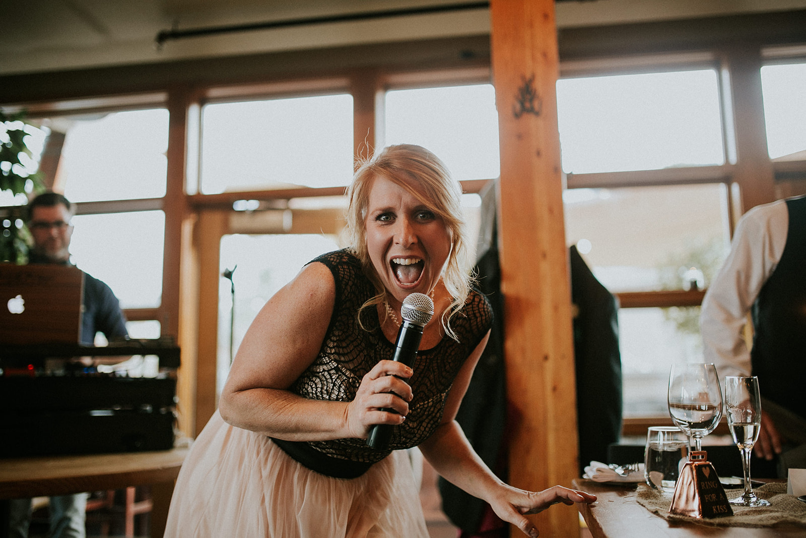 A woman with blonde hair in a sleeveless dress enthusiastically sings or speaks into a microphone, leaning forward at a wooden table set with glasses in a warmly lit room during a lively Banff wedding.
